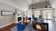 Located at 1160 Auston Grove Rd in Raleigh, NC, this single-floor clubhouse offers a well-appointed community gathering space designed for both comfort and functionality. The main lounge is a standout area, showcasing soaring vaulted ceilings, rich hardwood flooring, and a crisp white fireplace surround topped with a wall-mounted television — serving as a natural focal point for the room. Ceiling fans and abundant natural light filtering through multiple sets of French doors create an open, airy atmosphere, while neutral gray walls and bold blue accent furnishings lend the space a polished, contemporary feel. The lounge flows seamlessly into a dedicated dining area, which features classic crown molding, a tray ceiling detail, and a window pass-through that connects to the adjacent kitchen. The kitchen is equipped with wood cabinetry, stainless steel appliances, a bar-height counter with seating, and tile flooring — well-suited for hosting events or community gatherings. A separate, darker-toned room visible from the floor plan provides an additional flexible space. French doors throughout the clubhouse offer direct sightlines and access to outdoor areas, including a pool, reinforcing the bright, connected character of this community facility.