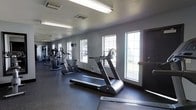 This fitness center is designed to provide a functional and comfortable environment for exercise. The space is laid out on a single floor, featuring a clean, open layout that accommodates a variety of cardio equipment, including treadmills, elliptical machines, and a stationary bike. The equipment is arranged in a linear fashion along one side of the room, allowing for easy access and an unobstructed workout area.

The interior is finished with neutral gray walls and durable dark rubber flooring, which is both practical and easy to maintain. Overhead fluorescent lighting runs the length of the ceiling, ensuring the space is consistently well-lit. A series of windows along the wall allows natural light to filter in, creating a bright and energizing atmosphere that enhances the overall workout experience.

The room provides enough open space to move comfortably between machines without feeling cramped. A wall-mounted television is also present, offering entertainment options during workouts. The overall ambiance is clean, modern, and purpose-driven, making it a practical fitness facility suited for daily use.