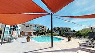 This outdoor amenity space is centered around a generously sized, geometric swimming pool with crystal-clear blue water, accessible via stainless steel entry rails. The expansive pool deck is finished in light-toned concrete pavers, providing ample room for lounging, and is lined with a collection of adjustable chaise lounge chairs arranged to take full advantage of the open sky above.

Striking terracotta-red triangular shade sails are suspended overhead at multiple points throughout the deck, offering stylish relief from direct sun while maintaining an open, airy feel. Decorative potted plants and ornamental grasses are placed around the pool's perimeter, adding greenery and visual interest to the hardscaped surroundings.

A dedicated fire pit lounge area is located adjacent to the pool, featuring a rectangular stone-surround fire feature filled with black lava rock, flanked by several cushioned black chairs. This seating area provides an inviting spot for relaxation both during the day and into the evening.

The entire space is enclosed by a dark metal fence, ensuring privacy and security while still allowing views of the surrounding landscaped grounds and multi-story residential buildings beyond. The overall ambiance is bright, open, and resort-inspired, designed to maximize outdoor enjoyment in a communal setting.