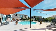 This outdoor amenity space is centered around a generously sized, geometric swimming pool with crystal-clear blue water, accessible via stainless steel entry rails. The expansive pool deck is finished in light-toned concrete pavers, providing ample room for lounging, and is lined with a collection of adjustable chaise lounge chairs arranged to take full advantage of the open sky above.

Striking terracotta-red triangular shade sails are suspended overhead at multiple points throughout the deck, offering stylish relief from direct sun while maintaining an open, airy feel. Decorative potted plants and ornamental grasses are placed around the pool's perimeter, adding greenery and visual interest to the hardscaped surroundings.

A dedicated fire pit lounge area is located adjacent to the pool, featuring a rectangular stone-surround fire feature filled with black lava rock, flanked by several cushioned black chairs. This seating area provides an inviting spot for relaxation both during the day and into the evening.

The entire space is enclosed by a dark metal fence, ensuring privacy and security while still allowing views of the surrounding landscaped grounds and multi-story residential buildings beyond. The overall ambiance is bright, open, and resort-inspired, designed to maximize outdoor enjoyment in a communal setting.