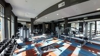 Located at 4625 Fairview Dr in Wilmington, NC, this fitness facility is housed on a single floor and offers a thoughtfully designed workout environment. The space features a striking curved architectural element along the ceiling, finished in a deep charcoal tone that contrasts cleanly with the bright white ceiling and recessed lighting above. Columns positioned throughout the floor plan provide structural definition while maintaining an open, airy feel. Large windows along the perimeter allow generous natural light to filter in, casting dynamic patterns across the floor and contributing to an energizing atmosphere. The flooring is a bold, patterned carpet in shades of blue, red, and white, adding a vibrant visual element to the space. The gym is outfitted with a comprehensive range of strength and cardio equipment, including cable machines, weight benches, rowing machines, and additional resistance training stations. Mirrored walls along multiple sides visually expand the space and support proper form during workouts. The overall ambiance is modern and motivating, combining sleek design details with functional fitness infrastructure to create a well-rounded exercise environment.