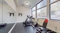 This dedicated fitness room offers a clean, functional layout designed to support a well-rounded workout routine. The space is finished with dark rubber flooring throughout, providing a durable and slip-resistant surface suitable for a variety of exercises. White walls and a neutral color palette create a bright, open atmosphere, while flush-mounted ceiling lights ensure consistent illumination across the room.

A standout feature is the full-length mirror wall that runs along one side of the space, visually expanding the room and allowing users to monitor their form during workouts. Large, floor-to-ceiling windows line the opposite wall, inviting ample natural light and providing views to the outside.

The room is equipped with a well-rounded selection of cardio and strength equipment, including an elliptical machine, a stationary bike, a treadmill, and an adjustable weight bench. A rack of dumbbells and a set of weight plates are neatly organized in one corner, complementing the wall-mounted pull-up and suspension training bars. A storage cabinet near the entry provides additional organizational space. The room is accessed through a secure door, making it a self-contained, purpose-built fitness facility.