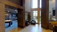 This striking space makes an immediate impression with its soaring double-height ceilings that flood the interior with an abundance of natural light. Expansive floor-to-ceiling windows frame views of a lush, landscaped exterior, seamlessly connecting the indoors with the greenery outside. The architectural design showcases rich, dark wood wall paneling that contrasts beautifully with the light hardwood flooring, creating a warm yet contemporary aesthetic throughout.

A carefully curated seating area anchors the main space, featuring two deep-cushioned dark leather armchairs arranged around a central ottoman, offering a comfortable and inviting place to gather. Decorative elements include a tall indoor tropical plant and a framed botanical artwork, adding organic texture and visual interest to the refined interior.

Adjacent to the main area, a secondary space is visible, equipped with what appears to be a monitor or screen display and additional furnishings, suggesting a functional or amenity area. Recessed and accent lighting complement the natural light, ensuring the space remains well-illuminated throughout the day. The overall ambiance is one of polished sophistication, blending modern architectural details with warm materials to create a welcoming and impressive environment.