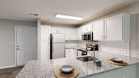 Located at 100 Patriot's Pointe Dr in Hillsborough, North Carolina, this single-floor residence offers a thoughtfully designed layout with modern finishes throughout. The open-concept kitchen is a standout feature, showcasing white shaker cabinetry, granite countertops, a classic subway tile backsplash, and a full suite of stainless steel appliances including a refrigerator, electric range, and built-in microwave. A peninsula with an undermount sink provides additional prep space and serves as a natural divider between the kitchen and the living area. The living room is generously proportioned, finished with neutral carpet, and equipped with a ceiling fan. Wood-look vinyl flooring flows through the entry, kitchen, and secondary spaces, adding warmth and continuity. The bedrooms offer comfortable proportions with built-in closet shelving for organized storage. The bathrooms are cleanly finished with white vanity cabinetry, light countertops, wood-look flooring, and a shower with a curtain enclosure. Neutral wall tones are carried throughout, creating a cohesive and move-in-ready aesthetic that suits a variety of furnishing styles.