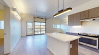 This contemporary single-floor unit showcases a thoughtfully designed open-concept layout that seamlessly integrates the living and kitchen areas. A striking glass-paneled garage door serves as a dramatic architectural feature in the main living space, flooding the area with natural light and offering outdoor views. Light wood-look flooring runs consistently throughout, enhancing the bright and airy atmosphere.

The kitchen is a standout, outfitted with sleek, wood-toned cabinetry extending to the ceiling, a crisp white waterfall-edge island with pendant lighting overhead, and a full suite of stainless steel appliances including a French door refrigerator, electric range, and dishwasher. Generous counter space and under-cabinet lighting add both function and style.

The bedroom is well-proportioned with a ceiling fan, a window with views of surrounding greenery, and light-toned flooring that continues the cohesive design language. A spacious walk-in closet with wrap-around hanging rods provides ample storage.

The bathroom features large-format tile flooring, a wood-accented vanity with a white countertop, a soaking tub with classic white subway tile surround, and a separate glass-enclosed shower. The overall palette is clean and neutral, creating a calm, modern ambiance throughout the space.