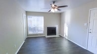 This single-floor residential space offers a clean, functional layout designed for comfortable everyday living. The living area serves as the centerpiece of the home, immediately drawing attention with its rich, dark hardwood flooring that runs throughout and provides a grounding contrast to the neutral, light-toned walls. A tile-surround fireplace anchors one wall, offering both a functional heating element and a classic architectural focal point that adds warmth and character to the space. Overhead, a ceiling fan with an integrated light fixture provides both air circulation and ambient lighting, making the room practical across all seasons. Natural light plays a significant role in the ambiance of this space — large windows dressed with horizontal blinds allow sunlight to pour in, creating a bright and airy atmosphere while still maintaining privacy control. The textured ceiling adds a subtle traditional touch to the overall design. The open feel of the living area, combined with the continuity of the flooring and the simplicity of the wall finishes, gives the space a versatile, move-in-ready quality that can accommodate a wide range of interior styles. Overall, this space balances practicality with comfort in a straightforward, well-maintained residential setting.