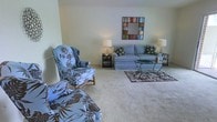 This single-floor space features a welcoming living area designed for comfort and functionality. The sitting area is anchored by a light blue upholstered sofa adorned with decorative floral and geometric accent pillows, complemented by a matching wingback armchair and ottoman upholstered in a bold tropical leaf print in shades of blue and brown. A glass-topped coffee table with an ornate metal base serves as a central focal point, while a coordinating side table with a table lamp provides warm ambient lighting. A second lamp on the opposite side of the sofa ensures balanced illumination throughout the space. The floors are covered in plush, neutral-toned carpeting, which adds warmth underfoot, and a patterned area rug defines the seating arrangement. A colorful mosaic-framed mirror mounted on the wall adds a decorative accent and visual interest. The walls are finished in a soft, neutral tone, contributing to an overall atmosphere that feels calm and inviting. The layout of the room is open and spacious, allowing for easy movement and a sense of airiness throughout the living area.