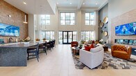 Located at 12631 Vance Jackson Rd in San Antonio, TX, this impressive amenity center spans a generously proportioned single floor, offering residents a comprehensive and upscale community experience. The entrance greets visitors with soaring double-height ceilings and dramatic floor-to-ceiling steel-framed glass doors that flood the space with natural light, setting an elegant and welcoming tone throughout.

The main lounge area features a striking accent wall clad in hexagonal mosaic tile, anchored by a sleek linear fireplace and a large mounted television — an ideal gathering space with refined furnishings and an airy ambiance. Adjacent seating nooks provide quieter retreats, adorned with vibrant artwork, large windows overlooking lush landscaping, and plush, contemporary furniture.

The fitness center is expansive and well-appointed, housing a full array of strength and cardio equipment, including treadmills, cycling bikes, stair climbers, and cable machines, all complemented by floor-to-ceiling mirrors and flat-screen televisions. Natural light pours in through wide windows framing views of the courtyard and palm trees.

Outdoors, a resort-style pool and spa serve as the centerpiece of the courtyard, surrounded by lounge chairs, umbrella-shaded seating, decorative fountains, and lush landscaping. The multi-story building architecture, featuring brick and stucco facades with private balconies, creates a vibrant and enclosed outdoor environment. A dedicated game room and utility room round out the functional offerings of this exceptional amenity space.