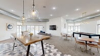 This thoughtfully designed single-floor communal space offers an open, airy layout that seamlessly blends multiple functional zones into one cohesive environment. The interior is anchored by a striking geometric hexagonal tile floor in the kitchen and lounge entry area, transitioning to plush carpeting and wood-look tile throughout the remaining zones.

The kitchenette is a standout feature, showcasing rich navy blue shaker-style cabinetry, crisp white countertops, open wood shelving, brass hardware, and stainless steel appliances — all illuminated by stylish pendant lights. A large butcher-block high-top communal table with acrylic barstools serves as the centerpiece of the kitchen area.

The lounge zone is anchored by a bold white shiplap accent wall with a wall-mounted flat-screen TV, complemented by a plush sectional sofa and blue accent chairs. Exposed industrial-style ductwork adds an urban, loft-inspired character to the ceiling.

A bright dining nook features a round white table with leather chairs, framed contemporary artwork, and oversized windows and sliding glass doors that flood the space with natural light and offer scenic outdoor views. A bay window area provides an additional casual seating nook. The space also includes a recreation area, completing this versatile and well-appointed gathering space.