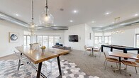 This thoughtfully designed single-floor communal space offers an open, airy layout that seamlessly blends multiple functional zones into one cohesive environment. The interior is anchored by a striking geometric hexagonal tile floor in the kitchen and lounge entry area, transitioning to plush carpeting and wood-look tile throughout the remaining zones.

The kitchenette is a standout feature, showcasing rich navy blue shaker-style cabinetry, crisp white countertops, open wood shelving, brass hardware, and stainless steel appliances — all illuminated by stylish pendant lights. A large butcher-block high-top communal table with acrylic barstools serves as the centerpiece of the kitchen area.

The lounge zone is anchored by a bold white shiplap accent wall with a wall-mounted flat-screen TV, complemented by a plush sectional sofa and blue accent chairs. Exposed industrial-style ductwork adds an urban, loft-inspired character to the ceiling.

A bright dining nook features a round white table with leather chairs, framed contemporary artwork, and oversized windows and sliding glass doors that flood the space with natural light and offer scenic outdoor views. A bay window area provides an additional casual seating nook. The space also includes a recreation area, completing this versatile and well-appointed gathering space.