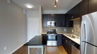 This single-floor space showcases a contemporary design with a well-appointed kitchen at its heart. The kitchen is outfitted with dark espresso cabinetry that extends to the ceiling, providing generous storage, and is complemented by light granite countertops that create a striking contrast. Stainless steel appliances, including a gas range, built-in microwave, and dishwasher, lend a polished, modern aesthetic. A textured mosaic tile backsplash adds visual depth and character to the cooking area, while a stainless steel undermount sink is positioned beneath a practical workspace. Warm hardwood flooring runs throughout, adding a natural element that balances the sleek, dark finishes. A compact dining area with a modern table sits just adjacent to the kitchen, making efficient use of the open layout. Track lighting overhead provides targeted illumination, while flush-mount fixtures contribute to an evenly lit environment. The white entry door features clean lines that complement the overall neutral color palette of the walls, contributing to a bright and airy atmosphere. This space is designed with functionality and modern style in mind, offering a cohesive and inviting living environment.