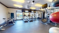 Located at 2550 W Ironwood Hill Dr in Tucson, Arizona, this fitness center is a comprehensive, single-floor facility designed to support a wide range of workout routines. The space is thoughtfully laid out across an open floor plan with durable rubber flooring throughout, providing both safety and comfort for exercise.

The cardio zone is prominently positioned along a wall of arched windows that flood the area with natural light and offer views of the outdoor pool area. This section is outfitted with multiple treadmills, elliptical trainers, and stationary spin bikes, catering to a variety of cardiovascular training preferences. Wall-mounted flat-screen TVs are positioned above the windows to keep members entertained during workouts.

The strength training area features a multi-functional cable and pulley machine system, a dedicated free weight section with a full rack of dumbbells, and mirrored walls that enhance the sense of space while aiding in proper form. Additional equipment includes rowing machines, a mini trampoline, and a weight bench, offering a diverse selection for full-body conditioning.

Overhead, multiple ceiling fans and recessed panel lighting ensure consistent air circulation and bright, even illumination. Crown molding along the upper walls adds a polished architectural touch. The facility is well-maintained and spacious, providing ample room to move freely between equipment zones.