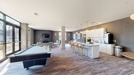 Located at 6300 Clayton Ave in St. Louis, Missouri, this single-floor amenity space offers a well-appointed, open-concept layout designed for both relaxation and socializing. The interior reflects a contemporary design aesthetic, with wood-look flooring, textured gray wall treatments, and a neutral color palette that creates a cohesive and inviting atmosphere throughout.

The lounge area serves as a central gathering point, anchored by a sleek gas fireplace set within a floor-to-ceiling dark tile surround. A wall-mounted television above the fireplace complements the seating arrangement, which includes a mid-century-inspired sofa, accent chairs, and a round ottoman. Floor-to-ceiling windows flood this area with natural light and provide views of the surrounding neighborhood.

The kitchen area is a standout feature, showcasing white cabinetry with subway tile backsplash, stainless steel appliances, and a large island topped with granite countertops. The island's decorative patterned tile base adds a distinctive design element, while globe pendant lights overhead enhance the modern aesthetic. Bar-height seating lines the island, making it ideal for casual dining or conversation.

A billiards table occupies a central position in the open layout, reinforcing the space's recreational focus. A banquette nook with pendant lighting and curated wall art provides a more intimate setting for smaller gatherings.