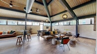 Located at 106 Kane St in West Hartford, CT, this single-floor community lounge is a thoughtfully designed gathering space that blends industrial architecture with warm, inviting décor. The centerpiece of the room is its dramatic vaulted ceiling, finished with natural wood planking and framed by bold dark green steel trusses that add both structural character and visual interest. Ceiling fans and globe pendant light fixtures hang from the pitched ceiling, enhancing both comfort and ambiance throughout the space.

Wide-plank hardwood flooring in a herringbone pattern runs throughout, complementing the earthy, mid-century modern aesthetic. A generous seating area anchors the main lounge, featuring a large leather sectional sofa, accent chairs, and a rustic wood coffee table set atop a patterned area rug. The recreational zone includes a full-size pool table and a foosball table, making the space well-suited for social gatherings. High-top bar tables with stools provide additional casual seating options.

A band of clerestory windows wraps around the upper perimeter of the walls, flooding the interior with natural light and offering views of the surrounding greenery. Textured wall tiles add a refined detail to the lower wall sections. A wall-mounted television and open shelving units round out the amenities, creating a versatile and dynamic communal environment.
