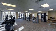 This fitness facility offers a thoughtfully designed workout environment spread across a single floor. The space is outfitted with a wide array of strength-training machines, including cable systems, chest press, leg press, and various selectorized weight machines, all arranged to allow for comfortable movement between stations. A dedicated cardio section features treadmills and upright bikes positioned near large floor-to-ceiling windows, allowing natural light to flood the space and providing views to the outdoors. A free weights area includes a dumbbell rack along a mirrored wall, catering to a range of workout preferences. The flooring throughout consists of durable, patterned carpet tiles well-suited for a high-traffic fitness environment. The ceiling features a suspended tile grid with recessed fluorescent panel lighting, ensuring the space is evenly and brightly lit. A glass-door entrance opens directly into the main workout floor, contributing to the open, accessible feel of the facility. The overall layout is clean, organized, and spacious, offering ample room between equipment to accommodate multiple users comfortably. This facility is well-suited for a full-body workout routine in a professional and functional setting.