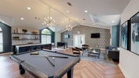 This thoughtfully designed clubhouse offers a welcoming and versatile gathering space across a single open floor. Soaring vaulted ceilings with dramatic tray detailing lend an airy, upscale character throughout, while herringbone-patterned wood-look flooring ties the various zones together cohesively.

The main game and social area is anchored by a full-size pool table and a foosball table, set beneath a striking sputnik chandelier. A bold geometric-patterned accent wall and wall-mounted televisions add visual interest and entertainment functionality to this zone. Adjacent seating areas feature plush sofas, velvet accent chairs, and ottomans arranged on area rugs, creating inviting spaces for relaxed conversation.

A cozy banquette nook provides an intimate retreat, framed by dark accent walls and illuminated by a sculptural cluster of ring pendant lights. A round table with upholstered chairs makes this corner ideal for small group gatherings.

The kitchen-style bar area is a standout feature, showcasing a large granite-topped island with gold-accented barstools, dark cabinetry, subway tile backsplash, and open shelving. Arched windows throughout the space flood the interior with natural light and offer views of the surrounding greenery, enhancing the overall ambiance with warmth and brightness.