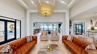 This single-floor space in Huntsville, Alabama offers a generously sized, open-concept layout designed for both social gatherings and focused work. The interior is unified by light hardwood flooring and crisp white walls, creating a bright, welcoming atmosphere throughout.

The main lounge area commands attention with dramatically soaring ceilings and a striking gold drum chandelier overhead. Tufted cognac leather sofas and neutral accent chairs define the seating arrangement, while large black-framed windows draw in abundant natural light and offer views of the surrounding community.

Adjacent to the lounge, a dedicated dining and meeting area features a substantial dark stone-topped communal table with curved chairs, illuminated by a brass rectangular pendant fixture. Bar-height bistro tables provide additional seating nearby, and a kitchenette with dark cabinetry, gold hardware, stainless steel appliances, and a sink serves the area conveniently.

A secondary lounge zone offers a more intimate setting, anchored by a linear fireplace set into a warm wood-paneled wall and accessed through sleek black-framed glass doors. A private office area features a bold geometric patterned accent wall, built-in workspace, and display surfaces. Throughout, curated artwork and decorative accents contribute to the space's contemporary, upscale character.
