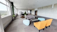 Located at 229 Andover Park E in Tukwila, Washington, this contemporary amenity floor offers a thoughtfully designed environment that blends comfort, function, and modern aesthetics. The open lounge area serves as the centerpiece of the space, featuring a mix of stylish seating including gray sofas, vibrant orange accent chairs, and sculptural light-blue ottomans arranged around sleek coffee tables. Exposed concrete accent walls add an industrial character that contrasts with the warmer soft furnishings, while living green walls and potted plants introduce natural elements throughout.

The fitness center is generously proportioned and well-equipped with a full array of cardio machines, including treadmills, ellipticals, and stationary bikes, as well as strength training equipment and a dumbbell rack. Large windows flood the gym with natural light and frame expansive views of the Tukwila skyline and surrounding greenery.

A modern kitchen features dark lower cabinetry paired with glossy white upper cabinets, a stainless steel refrigerator, microwave, and ample counter space, along with a central island. A secondary lounge area with soft neutral tones and broad windows further enhances the sense of openness, offering sweeping panoramic views. Neutral carpet tile flooring and drop-panel ceilings are consistent throughout, contributing to a clean, professional ambiance.
