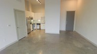 This single-floor space showcases a sleek, contemporary design with an open-concept layout that maximizes the sense of space and flow. The polished concrete floors throughout lend an industrial-chic aesthetic, reflecting light and adding a refined yet minimalist character to the interior.

The kitchen is a standout feature, outfitted with white cabinetry, a classic subway tile backsplash, and a full suite of stainless steel appliances including a refrigerator, microwave, electric range, and dishwasher. Track lighting above the kitchen provides focused illumination, complementing the clean, modern feel of the space.

Soaring ceilings amplify the open atmosphere, creating an airy environment throughout the main living area. A structural column serves as a subtle architectural divider between the kitchen and the broader living space without disrupting the open-plan design. Two entry points are visible — one leading to an adjacent room or utility space and a secondary door that likely serves as the main entrance — offering a practical and well-organized layout. The neutral white walls and minimal design palette create a versatile canvas, well-suited for a variety of interior styles and uses.