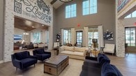 This single-floor community lounge is a thoughtfully designed social space that blends industrial character with modern comfort. The centerpiece of the space is its dramatic vaulted ceiling, which soars overhead and is clad in warm wood planking in select areas, creating an inviting, lodge-like atmosphere. Whitewashed brick columns punctuate the open floor plan, adding architectural texture and anchoring the various seating zones below.

The main lounge area features plush navy blue sofas and armchairs arranged around a rustic wood-and-metal coffee table, complemented by a large woven area rug and wide-plank hardwood flooring throughout. Floor-to-ceiling windows and multiple sets of glass French doors flood the interior with natural light and provide views of the surrounding exterior. A cozy fireplace nook with a wall-mounted television and blue accent chairs offers a more intimate gathering spot.

A fully equipped kitchenette lines one wall with dark cabinetry, open shelving, stainless steel appliances including a wall oven and refrigerator, and a clean white countertop. A separate dining area with a round table and upholstered chairs sits adjacent to the lounge. Distinctive hand-painted murals and custom signage — including a prominent "Free Roam Lounge" and "Social" artwork — lend the space a spirited, community-focused identity. The overall ambiance is warm, welcoming, and well-appointed.