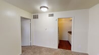This single-floor unit offers a straightforward and functional layout designed for comfortable everyday living. The main living area is finished with wall-to-wall carpet and features neutral cream-toned walls, a ceiling light fixture, and HVAC vents for climate control. A closet is accessible directly from the living area, adding convenient storage. Adjacent to the living space, the compact kitchen is equipped with essential appliances including an electric coil range with an overhead ventilation hood and a full-size refrigerator. White cabinetry lines the upper walls, providing ample storage, while rich hardwood-style flooring runs throughout the kitchen and connecting areas, offering a warm contrast to the carpeted rooms. The bathroom is thoughtfully appointed with a granite-style countertop vanity, an inset mirror with overhead lighting, a white toilet, built-in overhead cabinetry, and a full-size tub and shower combination with tiled surround. The bedroom is a bright, open room featuring soft carpeting, cream walls, and a window that allows natural light to filter in, creating a calm and restful atmosphere. A second window in the living area further enhances the sense of openness throughout the unit. Clean lines and a neutral palette give the space a simple, move-in-ready character.