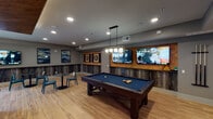 Located at 14175 Dallas Pkwy in Farmers Branch, Texas, this single-floor amenity lounge is a thoughtfully designed social and recreation space that blends contemporary style with entertainment-focused functionality. The space is unified by warm wood-look flooring that runs throughout, complemented by gray walls accented with textured, dark wood-grain wainscoting panels that lend a sophisticated, modern edge to the interior.

The game room serves as a central highlight, anchored by a full-size pool table with a deep blue felt top, suspended beneath a striking geometric pendant light fixture. A wood-paneled feature wall hosts multiple large flat-screen TVs and a built-in cue rack, making it equally suited for game nights and sports viewing. A dedicated dart area features two regulation dartboards framed within a warm wood surround, flanked by vivid abstract wall art, with bistro-style tables nearby for casual seating.

Adjacent to the game room, a lounge area offers a comfortable retreat centered around a plush neutral sofa and a live-edge coffee table. The focal wall here is a bold geometric design combining dark wood, natural wood, and gold metallic accents framing a mounted TV — a standout architectural detail. Recessed lighting throughout maintains a warm, inviting ambiance, while a marble-topped counter and glimpses of additional seating nooks complete this well-appointed communal space.