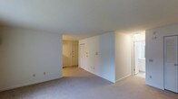 This single-floor residential space offers a straightforward and functional layout designed for comfortable everyday living. The main living area is finished with neutral-toned carpeting that extends throughout, contributing to a warm and cohesive feel across the space. The walls are painted in a clean, bright white, which helps maximize the sense of openness and reflects the available light effectively.

The layout features a central partial wall that creates a natural division between the main living area and an adjoining hallway or secondary space, offering a sense of structure without feeling overly segmented. A sliding closet is visible along this dividing wall, providing convenient built-in storage. The textured ceiling adds a subtle design detail consistent throughout the unit.

Doorways lead to additional areas of the home, including what appears to be an entry or utility space to one side and a hallway corridor on the other, suggesting a thoughtful flow between the functional zones of the residence. Ceiling-mounted lighting provides even illumination in the connecting areas.

Overall, the space presents a clean, move-in ready environment with a neutral palette, practical storage solutions, and a well-organized single-level floor plan that offers ease of navigation and a calm, uncluttered ambiance.