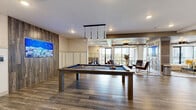 Located at 2450 Cherry Laurel Dr in Sanford, FL, this modern single-floor amenity lounge is thoughtfully designed to accommodate both social gatherings and relaxed lounging. The space is unified by wood-plank tile flooring that runs throughout, complemented by a mix of herringbone and linear patterns that add visual interest underfoot.

At the heart of the space sits a full-size pool table beneath elegant pendant lighting, serving as a central focal point for recreational activity. Adjacent to the game area, a dedicated lounge zone features plush seating arranged around a central coffee table, anchored by a decoratively paneled column with an embedded flat-screen display. A striking wood-paneled ceiling treatment adds warmth to this seating area.

A separate dining nook offers booth-style seating with navy upholstered benches, crushed-velvet accent chairs, and individual mounted televisions at each table, providing an intimate setting for casual dining or work. Floor-to-ceiling sliding glass doors and expansive windows flood the space with natural light and open to a railed outdoor balcony with community views. Wall-mounted artwork and curated décor complete the polished, contemporary ambiance throughout.
