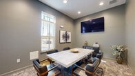 Located at 5401 Commonwealth Centre Pkwy in Midlothian, Virginia, this professional conference room offers a polished and modern setting well-suited for focused meetings and collaborative sessions. The room is finished with cool gray walls that lend a sophisticated, contemporary feel throughout the space.

At the center of the room sits a substantial concrete-top conference table surrounded by dark leather armchairs with warm wood-toned frames, providing comfortable seating for up to six participants. A large wall-mounted flat-screen display is positioned prominently on one wall, making it ideal for presentations and video conferencing.

Natural light enters through double-hung windows fitted with horizontal blinds, offering both brightness and privacy control. Recessed ceiling lighting supplements the natural light, ensuring the room remains well-lit at all times. A patterned area rug anchors the seating arrangement, adding texture and visual warmth to the otherwise clean-lined interior.

Decorative touches include botanical-inspired framed artwork on the walls and tasteful accent pieces atop a dark storage credenza. A frosted glass sliding door provides a semi-private entry point, adding an architectural element that enhances the room's refined aesthetic. The overall ambiance is professional, comfortable, and thoughtfully designed for productive use.
