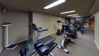 This Community Room and Fitness Center offers a versatile, single-floor amenity space designed to accommodate both recreation and wellness. The layout is divided into distinct zones, each serving a unique purpose for residents and guests.

The game area is a central highlight, featuring a full-size pool table with a bright blue felt surface and a foosball table, both set on neutral carpeted flooring beneath overhead fluorescent lighting. A decorative wallpaper border runs along the upper walls, adding a touch of character throughout the space.

Adjacent to the game area is a comfortable lounge, anchored by a classic brick fireplace that serves as the room's focal point. The seating area includes a sofa, armchairs, and a wooden coffee table, all arranged around the hearth. A wall-mounted television and a water cooler add convenience, while recessed lighting and a textured ceiling contribute to the warm, inviting ambiance.

The fitness center is well-appointed with a variety of cardio equipment, including treadmills, stationary bikes, and elliptical machines, along with strength training equipment. The room is brightly lit with overhead fluorescent panels and features the same carpeted flooring found throughout. A Pepsi vending machine and staircase access round out this fully functional amenity space.