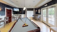 This sophisticated game room is designed for both entertainment and relaxation, centered around a full-size pool table with a slate-gray felt surface and rich mahogany wood finish, set atop a patterned area rug over light hardwood flooring. The room's distinctive aesthetic is defined by dramatic dark textured walls paired with crisp white wainscoting, creating a refined, club-like atmosphere. Recessed lighting and elegant pendant fixtures illuminate the space, while wall-mounted sconces add a warm, ambient glow. Multiple bar-height bistro tables with cushioned counter stools are arranged throughout the room, offering comfortable perches for spectators or casual conversation. A wall-mounted flat-screen television is positioned for easy viewing from multiple angles, making the space equally suited for watching sports or entertainment. Floor-length curtains frame large windows fitted with blinds, allowing for adjustable natural light, while a set of French doors opens to an outdoor area, inviting fresh air and greenery into the room. Contemporary framed artwork adorns the walls, complementing the space's polished, modern-classic design. An open doorway connects this room to an adjacent lounge and dining area, enhancing the social flow of the overall space.