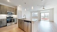 This thoughtfully designed one-bedroom, one-bathroom residence offers a modern and functional layout across a single floor. The open-concept kitchen serves as the centerpiece of the living area, showcasing wood-toned flat-front cabinetry extending to the ceiling for ample storage, a subway tile backsplash, and a full suite of stainless steel appliances including a range, microwave, and French door refrigerator with ice dispenser. A granite-topped island with an undermount sink and pendant lighting above provides additional prep space and a natural gathering point. Light wood-style flooring flows throughout the kitchen and living areas, complementing the clean white walls and recessed lighting overhead. A ceiling fan in the living area adds comfort and airflow. The bedroom is generously sized, featuring soft carpet underfoot, a ceiling fan, and a large window with exterior views that fills the room with natural light. The full bathroom is finished with large-format tile flooring, a granite-topped vanity with an illuminated mirror, and a soaking tub with tiled surround. A dedicated walk-in closet with built-in shelving and hanging rods is accessible directly from the bathroom, offering organized and convenient storage throughout.