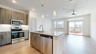 This thoughtfully designed one-bedroom, one-bathroom residence offers a modern and functional layout across a single floor. The open-concept kitchen serves as the centerpiece of the living area, showcasing wood-toned flat-front cabinetry extending to the ceiling for ample storage, a subway tile backsplash, and a full suite of stainless steel appliances including a range, microwave, and French door refrigerator with ice dispenser. A granite-topped island with an undermount sink and pendant lighting above provides additional prep space and a natural gathering point. Light wood-style flooring flows throughout the kitchen and living areas, complementing the clean white walls and recessed lighting overhead. A ceiling fan in the living area adds comfort and airflow. The bedroom is generously sized, featuring soft carpet underfoot, a ceiling fan, and a large window with exterior views that fills the room with natural light. The full bathroom is finished with large-format tile flooring, a granite-topped vanity with an illuminated mirror, and a soaking tub with tiled surround. A dedicated walk-in closet with built-in shelving and hanging rods is accessible directly from the bathroom, offering organized and convenient storage throughout.