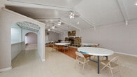 Located at 7575 Bissonnet St in Houston, TX, this single-floor gathering space offers a generous, open layout well-suited for events, meetings, or community functions. The main hall is defined by vaulted ceilings with exposed white-painted beams and a textured finish, lending the space a sense of volume and architectural character. Multiple ceiling fans are evenly distributed throughout, ensuring comfortable airflow across the entire area.

The most striking architectural feature is a floor-to-ceiling rustic stone fireplace, constructed from natural river rock, which serves as a commanding focal point along one wall. A raised stone hearth with a white railing detail adds depth and visual interest to the surrounding space. The flooring transitions between light vinyl tile and a warm terracotta-toned section near the fireplace, subtly defining gathering zones within the open floor plan.

Decorative arched openings with white columns frame the entry to a hallway, adding a refined architectural touch. The hallway connects to an adjacent kitchen area equipped with a stove, visible through the arched passageway. Windows along the perimeter allow natural light to filter in, brightening the neutral white and beige interior. The overall ambiance is functional yet characterful, combining rustic and traditional design elements in a versatile, open setting.