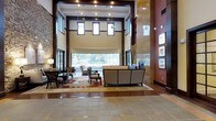 This impressive lobby showcases a dramatic double-height ceiling that immediately establishes a sense of grandeur and openness. Suspended pendant lighting fixtures hang at varying heights from the towering ceiling, complementing the recessed lighting that illuminates the space evenly throughout. A striking combination of materials defines the interior aesthetic, including exposed stone accent walls, rich dark wood trim, and warm beige plaster walls that together create a sophisticated yet inviting ambiance.

The seating area serves as the focal point of the space, featuring a generously sized sofa and coordinating armchairs arranged atop a large decorative area rug with a classic pattern. Additional seating clusters are positioned nearby, offering flexible arrangements for conversation or relaxation. Large windows along the rear wall flood the interior with natural light while framing views of the surrounding greenery outside.

The flooring transitions between polished hardwood in the seating area and large-format stone tile toward the entrance, adding visual depth and clear zone definition. Framed artwork lines the right accent wall, contributing a gallery-like quality to the space. Overall, the design balances functionality with refined, hotel-inspired elegance.