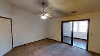 This single-floor space offers a straightforward and functional layout with neutral, easy-to-customize finishes throughout. The main living area is finished with wall-to-wall carpeting in a neutral tone, providing a comfortable underfoot feel and a clean, understated aesthetic. Walls are painted in a light, neutral hue, creating a bright and open atmosphere that can adapt to a variety of interior styles. A ceiling fan with an integrated light fixture is centrally mounted overhead, offering both air circulation and ambient lighting to the room. Dark-trimmed baseboards and door frames provide a subtle contrast against the light walls, adding a touch of visual definition to the space. A set of dark-framed sliding glass doors is a notable feature, opening into an adjoining room or enclosed area that benefits from natural light streaming through a visible window. This transition point enhances the sense of connectivity within the floor plan. A ceiling vent ensures climate control throughout the space. Overall, the layout is clean and versatile, well-suited for a range of uses and personal touches.