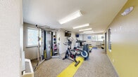 Located at 645 Wolff St in Denver, Colorado, this fitness center offers a dedicated exercise room designed to support a well-rounded workout routine. The space features a neutral color palette with light gray walls accented by bold yellow floor strips, giving it a modern, energetic feel. Flooring consists of low-pile carpet with yellow accent runners delineating the primary workout zones.

The room is equipped with a variety of cardio machines, including a Life Fitness treadmill, an elliptical trainer, and a stationary spin bike. A multi-station cable machine anchors one end of the room, accompanied by a rack of medicine balls in varying weights for functional training. Additional equipment, including a weight bench and recumbent bike, round out the offerings.

Two wall-mounted flat-screen TVs are positioned throughout the room, allowing users to stay entertained during workouts. Overhead fluorescent lighting ensures the space is consistently and brightly lit. Multiple windows line the walls, bringing in natural light and providing views of the surrounding outdoor area. A dedicated entrance door with secured access provides convenient entry directly from the exterior. Interior pass-through windows connect the fitness center to an adjacent common area, adding a sense of openness to the overall layout.