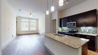 This single-story residence is designed with an open-concept layout that seamlessly connects the kitchen and living areas, creating a spacious and functional flow throughout. The kitchen is a standout feature, outfitted with rich dark cabinetry, granite countertops, a mosaic tile backsplash, and a full suite of stainless steel appliances including a refrigerator, range, and microwave. A central island with a granite surface and an integrated sink provides additional workspace and serves as a natural gathering point. Pendant lighting above the island and track lighting throughout add a polished, modern touch. Hardwood-style flooring extends through the main living areas, while large windows allow natural light to fill the space and offer views of the surrounding greenery. The living area is complemented by a ceiling fan for added comfort. Bedrooms are carpeted and feature soaring ceilings, generous closet space with built-in shelving, and access to natural light. The bathroom includes large-format tile flooring, dark vanity cabinetry with granite countertops, and a framed mirror. A dedicated laundry area accommodates a full-size washer and dryer, adding everyday convenience.
