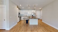 Located at 9235 Senator Royall Dr. in Charlotte, NC, this single-floor residence offers a thoughtfully designed layout with modern finishes and an open, airy feel throughout. The kitchen is a standout feature, showcasing crisp white shaker cabinetry, dark stone countertops, a classic subway tile backsplash, and a full suite of stainless steel appliances including a range, dishwasher, and French door refrigerator. A center island with a matching dark countertop provides additional prep space and casual seating, while pendant lighting adds a refined touch overhead. Warm, light-toned hardwood-style flooring flows seamlessly through the kitchen and into the adjacent living and dining areas, which are bathed in natural light from large windows. A glass-panel door in the living area leads directly outside, enhancing the connection to the outdoors. The bedroom features a ceiling fan and a generous walk-in closet outfitted with wire shelving for ample storage. The bathroom is well-appointed with a dual-sink vanity, dark stone countertops, white cabinetry with brushed nickel hardware, and framed mirrors. The overall ambiance is clean, contemporary, and move-in ready, reflecting a cohesive design sensibility throughout.