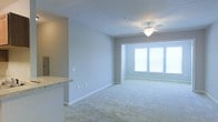 This single-floor residence offers a clean, modern aesthetic defined by an open-concept floor plan that encourages a natural flow between living and kitchen areas. The living space is finished with plush wall-to-wall carpeting in a warm neutral tone, providing a comfortable and inviting atmosphere. Soft gray walls complement the light color palette throughout, creating a calm and cohesive environment. A ceiling fan is centrally positioned in the living area, adding both functionality and a subtle design detail to the space. A large window along the far wall allows generous natural light to fill the room, enhancing the sense of openness and airiness. White horizontal blinds on the window offer privacy while still permitting diffused light to filter through. The kitchen area features a raised countertop with a light stone-pattern surface, providing a convenient breakfast bar that visually separates the kitchen from the living space without disrupting the open feel. Crisp white baseboards and trim run throughout, adding a polished finishing touch. Overall, the space presents a well-maintained, move-in-ready layout with a bright and neutral design that offers a versatile foundation for a variety of interior styles.