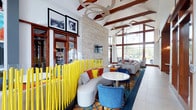 This single-story communal space showcases a bold, contemporary design centered around a dramatic double-height lobby area. Soaring vaulted ceilings with exposed wood beams are complemented by sculptural ring-shaped pendant lighting, creating an eye-catching architectural focal point overhead. Floor-to-ceiling windows along the perimeter flood the interior with abundant natural light while framing outdoor views.

The main lounge area features a striking stacked-stone accent wall housing a linear gas fireplace with a mounted flat-screen television above. Seating is arranged thoughtfully throughout, including curved gray sofas with colorful accent pillows, deep blue velvet chairs, and white pedestal tables, all anchored by a vivid abstract-patterned area rug in blue, orange, and gold tones.

A distinctive yellow vertical rod partition defines a cozy banquette seating zone, adding a playful sculptural element to the space. A separate intimate nook features curved, floor-to-ceiling burnt-orange upholstered walls with built-in seating and a globe pendant light fixture, offering a private retreat. A dedicated seating area near the entry provides additional relaxation space alongside a built-in coffee and beverage station, enhancing the space's functionality and welcoming ambiance.