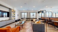 Located at 346 W 26th St in Charlotte, NC, this single-floor community amenity space offers a thoughtfully designed open-plan layout spanning a generous footprint. The interior is organized into several distinct functional zones that flow seamlessly into one another, all unified by light hardwood-style flooring, recessed lighting, and crisp white walls.

The main lounge area features multiple seating arrangements, including a large tufted leather sectional, an accent armchair in bold orange, and a curved caramel leather banquette beneath a statement globe chandelier — ideal for both casual gatherings and social events. Built-in shelving with integrated lighting frames a wall-mounted television, providing a focal point for the entertainment zone.

A dedicated kitchen and bar area is equipped with sage-green cabinetry, granite and dark stone countertops, mirrored subway tile backsplash, under-counter beverage refrigerators, and pendant lighting in an industrial-inspired finish. A large central island anchors the space for both food prep and informal dining.

Additional seating nooks — including a sunlit green tufted sectional near double glass doors — offer quiet retreat options. Large windows throughout invite ample natural light and provide views of the surrounding Charlotte neighborhood, lending the entire space an open, welcoming ambiance.