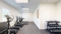 Located at 5095 Big Lake Rd in Lake Charles, Louisiana, this fitness center offers a clean, well-organized exercise space designed to support a variety of cardio-focused workouts. The room is finished with crisp white walls and durable rubber flooring, creating a practical and hygienic environment suited for regular physical activity.

The exercise room is equipped with multiple True-brand machines, including treadmills, elliptical trainers, and a recumbent stationary bike, providing users with diverse cardio options. A weight bench is also present, adding a strength-training element to the space. The equipment is arranged efficiently along the perimeter and interior of the room, allowing for comfortable movement between machines.

Natural light enters through several windows fitted with horizontal blinds, offering views of the surrounding greenery and contributing to an open, airy atmosphere. Overhead, flush panel skylights and recessed ceiling vents supplement the lighting and ventilation throughout the space. A direct exterior door provides convenient access to and from the outdoors.

The layout is thoughtfully organized, with open floor space remaining for stretching or additional use. A wall-mounted panel and pass-through window suggest potential for administrative or utility functions. Overall, the fitness center presents a functional and well-maintained environment for residents or users seeking accessible exercise facilities in Lake Charles.