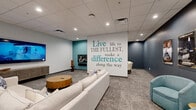 Located at 355 N Rosalind Ave in Orlando, FL, this single-floor lounge and entertainment space offers a well-appointed interior designed for comfort, collaboration, and casual gatherings. The layout is open and generously sized, accommodating multiple distinct zones within a single interconnected floor plan.

The space is anchored by a central lounge area featuring a large, plush sectional sofa paired with a round wooden coffee table and a wall-mounted large-format television above a slatted wood media console. An eye-catching accent wall displays the motivational phrase "Live life to the fullest, make a difference along the way" in teal and navy lettering, setting an energetic tone throughout.

A secondary seating zone features a round game or conference table surrounded by upholstered chairs, ideal for group activities or meetings. Two teal barrel chairs provide additional casual seating along a deep navy accent wall adorned with a curated gallery of classic film posters.

The kitchenette area includes warm wood cabinetry, a double-basin sink with a modern faucet, a countertop refrigerator, and floating shelves styled with decorative accessories. Recessed lighting throughout the drop-panel ceiling ensures consistent, even illumination across all areas. Neutral patterned carpet ties the various zones together, creating a cohesive and professional yet inviting atmosphere.