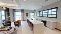 Located in Corinth, Texas, this single-floor community space is thoughtfully designed to blend contemporary aesthetics with warm, inviting elements. The open layout is anchored by a spacious lounge area featuring a striking reclaimed wood accent wall with a wall-mounted flat-screen television. Comfortable upholstered armchairs are arranged around a central coffee table, creating an ideal setting for relaxed gatherings or casual conversation. Ambient lighting from a tray ceiling adds a soft, refined glow to the lounge.

Adjacent to the seating area is a well-appointed kitchenette, highlighted by an expansive white quartz island with bar seating for multiple guests. The counter space along the far wall is fitted with modern grey cabinetry, an undermount sink, a built-in beverage refrigerator, and pendant lighting overhead. A wide bank of dark-framed windows runs the length of the back wall, framing sweeping views of the surrounding landscape and flooding the space with natural light.

Large sliding glass doors open to outdoor views, further enhancing the bright, airy atmosphere. Light-toned large-format tile flooring unifies the space, complementing the clean, neutral color palette throughout. An elevator provides accessibility, and the overall design strikes a balance between functional and sophisticated, making it a versatile space for community use.