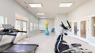 Located in Kathleen, Georgia, this fitness center offers a clean, modern exercise space designed to support a well-rounded workout routine. The single-floor layout is thoughtfully organized to maximize functionality while maintaining an open, uncluttered feel.

The exercise room features light-toned hardwood-style flooring and crisp white walls that lend a bright, fresh atmosphere throughout. A series of large recessed panel skylights line the ceiling, flooding the space with abundant natural light and contributing to the room's airy, energizing ambiance. Windows with shutters along the walls further enhance ventilation and visibility.

Cardio equipment is prominently featured, with multiple Spirit-brand treadmills and elliptical machines arranged for easy access and comfortable use. A dedicated free weight and strength area includes adjustable dumbbells and a workout bench, while a colorful stability ball rack adds a versatile functional training component. A wall-mounted water fountain provides added convenience for users during workouts.

The layout allows ample circulation space between equipment, ensuring a safe and comfortable experience. A pass-through window connects to an adjacent area, offering a sense of openness. The exterior entrance features a glass-panel door providing views to the outdoors. Overall, the fitness center presents a welcoming, well-maintained environment suited for a variety of exercise routines.