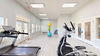 Located in Kathleen, Georgia, this fitness center offers a clean, modern exercise space designed to support a well-rounded workout routine. The single-floor layout is thoughtfully organized to maximize functionality while maintaining an open, uncluttered feel.

The exercise room features light-toned hardwood-style flooring and crisp white walls that lend a bright, fresh atmosphere throughout. A series of large recessed panel skylights line the ceiling, flooding the space with abundant natural light and contributing to the room's airy, energizing ambiance. Windows with shutters along the walls further enhance ventilation and visibility.

Cardio equipment is prominently featured, with multiple Spirit-brand treadmills and elliptical machines arranged for easy access and comfortable use. A dedicated free weight and strength area includes adjustable dumbbells and a workout bench, while a colorful stability ball rack adds a versatile functional training component. A wall-mounted water fountain provides added convenience for users during workouts.

The layout allows ample circulation space between equipment, ensuring a safe and comfortable experience. A pass-through window connects to an adjacent area, offering a sense of openness. The exterior entrance features a glass-panel door providing views to the outdoors. Overall, the fitness center presents a welcoming, well-maintained environment suited for a variety of exercise routines.