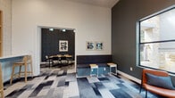 This thoughtfully designed single-floor space functions as a multifunctional lounge and co-working area, combining social and productive zones within a cohesive, modern aesthetic. Soaring ceilings amplify the sense of openness throughout, while large black-framed windows invite abundant natural light and offer views of an outdoor courtyard lined with stone architecture and native plantings.

The main lounge area features a curated seating arrangement with cognac leather chairs, a teal upholstered ottoman, and a built-in banquette accented by small side tables. Framed abstract artwork adorns the dark charcoal accent walls, adding visual interest against the crisp white upper walls. A geometric, multi-toned carpet in shades of blue, grey, and white anchors the space and carries consistently throughout.

A built-in counter with bar-height seating and open wood shelving provides a casual co-working station, decorated with plants and decorative objects. The patterned tile backsplash behind the shelving adds texture and warmth to this work nook.

An adjacent meeting room, separated by a glass-paneled sliding door, features dark textured wallcovering, a rectangular wood table, and surrounding chairs — ideal for small group gatherings or focused work sessions. Together, the zones create a versatile, design-forward environment suited for both collaboration and relaxation.