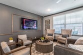 This single-story community space is defined by a dramatic entry corridor anchored by soaring vaulted ceilings with exposed natural wood beams, creating an immediate architectural statement. Large-format light stone tile flooring runs the length of the hallway, while a decorative console table and curated artwork add a polished, welcoming ambiance. Glass-paneled doors flank the corridor, allowing natural light to filter through and providing views to the exterior.

The layout opens into multiple distinct gathering areas. A primary lounge features a large wall-mounted television, a mix of tufted wingback chairs, a sectional sofa, and a herringbone-patterned wood-look floor, anchored by a bold geometric area rug. A separate, more intimate media nook offers a sectional sofa, wall-mounted TV, and contemporary artwork for a cozy retreat. An additional bistro-style seating area with a round wood table and tufted chairs sits beside large windows, providing natural light and outdoor views.

The fully equipped kitchen includes stainless steel appliances, a gas range, subway tile backsplash, white shaker-style cabinetry, gray countertops, and herringbone flooring—offering ample functionality for communal use. The 3D floor plan reveals a billiards table, further reinforcing the space's role as a comprehensive social hub.