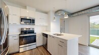 This contemporary single-floor unit offers a well-designed layout that balances modern aesthetics with functional living. The open-concept kitchen is a standout feature, equipped with stainless steel appliances including a range, microwave, and dishwasher. Crisp white upper cabinetry contrasts with light wood-toned lower cabinets, while a quartz waterfall peninsula with an undermount sink and gold-accented pendant lighting adds a refined touch. Exposed metallic ductwork running along the ceiling lends an industrial character to the space.

The living area is bright and open, benefiting from large sliding glass doors that flood the room with natural light and provide direct access to a generously sized private outdoor terrace. The terrace is finished with artificial turf and stone pavers, enclosed by corrugated metal privacy panels, offering a unique outdoor retreat with open sky views.

The bedroom is calm and neutral, featuring durable wood-look flooring and a spacious walk-in closet with built-in shelving. The bathroom is elegantly appointed with large-format marble-look tile on both the floors and shower walls, a frameless sliding glass shower enclosure, and clean, minimalist fixtures throughout. Wood-look flooring is consistent across the main living areas, tying the space together cohesively.